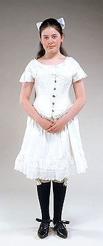 girl wearing wide white hair ribbon tied in a bow at the back of her head, white cotton corset over a white cotton slip and black shoes that tie with a bow over the instep.