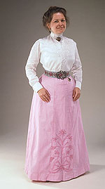 woman wearing long-sleeved white cotton shirt with a high collar and pin at the throat and a floor-length pink skirt.